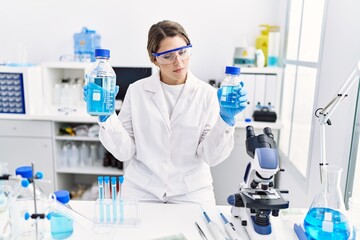 Young hispanic woman wearing scientist uniform holding liquid bottle at laboratory