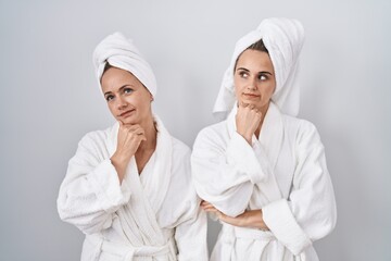 Middle age woman and daughter wearing white bathrobe and towel with hand on chin thinking about question, pensive expression. smiling with thoughtful face. doubt concept. - obrazy, fototapety, plakaty