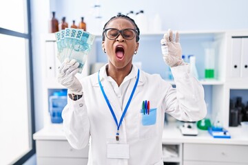 African woman with braids working at scientist laboratory holding money angry and mad screaming frustrated and furious, shouting with anger looking up.