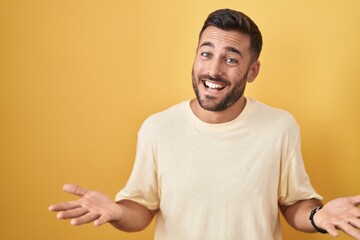 Handsome hispanic man standing over yellow background smiling cheerful with open arms as friendly welcome, positive and confident greetings