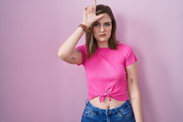 Blonde caucasian woman standing over pink background making fun of people with fingers on forehead...