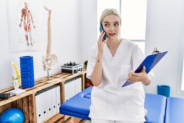 Young caucasian woman wearing physio therapist uniform talking on the smartphone at clinic