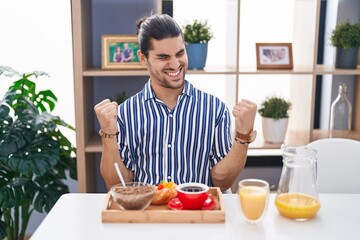 Fototapeta na wymiar Hispanic man with long hair sitting on the table having breakfast very happy and excited doing winner gesture with arms raised, smiling and screaming for success. celebration concept.