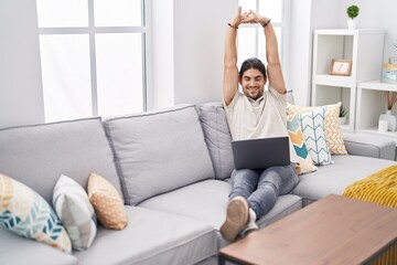 Young hispanic man using laptop stretching arms at home