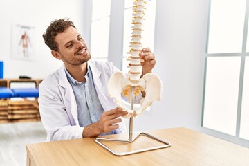 Young hispanic man wearing physiotherapist uniform touching anatomical model of vertebral column at...