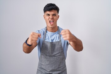 Hispanic young man wearing apron over white background angry and mad raising fists frustrated and furious while shouting with anger. rage and aggressive concept.