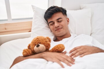 Young hispanic man hugging teddy bear lying on bed sleeping at bedroom