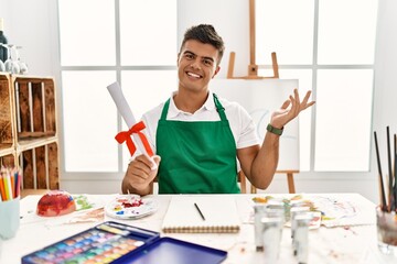 Young hispanic man at art studio holding degree smiling cheerful presenting and pointing with palm of hand looking at the camera.