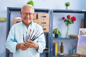 Senior grey-haired man artist smiling confident holding paintbrushes at art studio