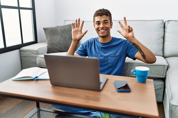 Young handsome hispanic man using laptop sitting on the floor showing and pointing up with fingers number eight while smiling confident and happy.