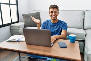 Young handsome hispanic man using laptop sitting on the floor smiling cheerful presenting and pointing with palm of hand looking at the camera.