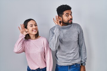 Young hispanic couple standing together smiling with hand over ear listening an hearing to rumor or gossip. deafness concept.