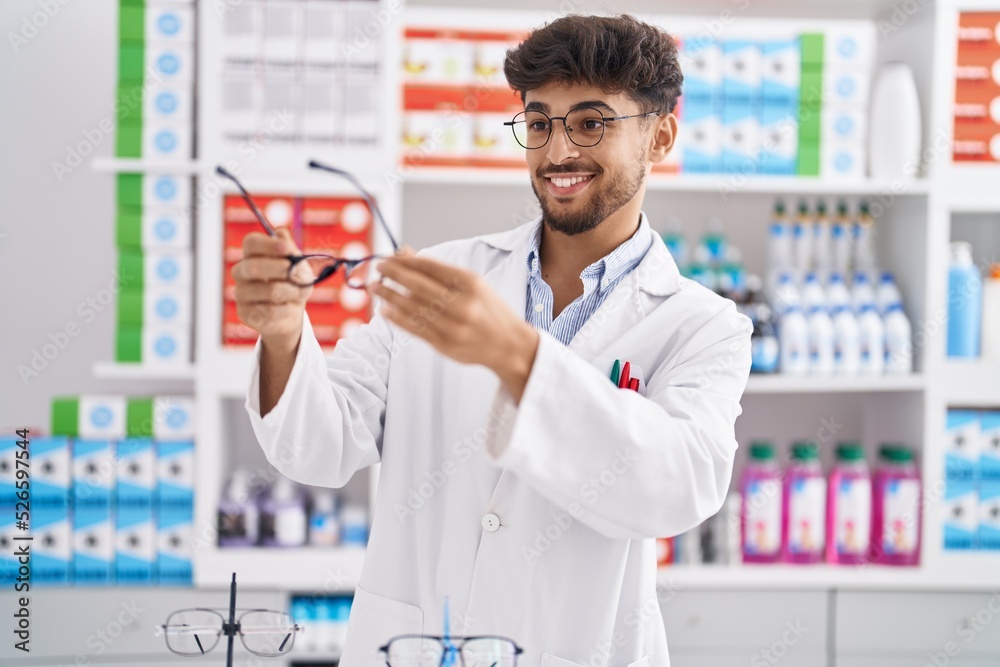 Poster Young arab man pharmacist smiling confident holding glasses at pharmacy