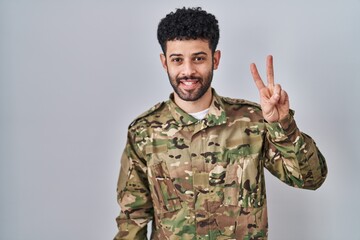 Arab man wearing camouflage army uniform showing and pointing up with fingers number two while smiling confident and happy.