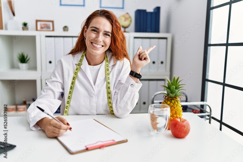 Wall mural Young redhead woman nutritionist doctor at the clinic with a big smile on face, pointing with hand finger to the side looking at the camera.