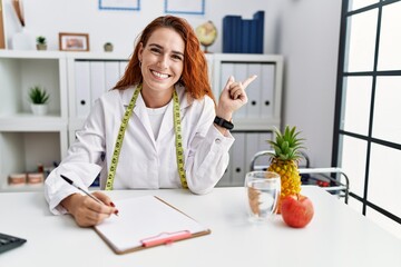 Young redhead woman nutritionist doctor at the clinic with a big smile on face, pointing with hand...