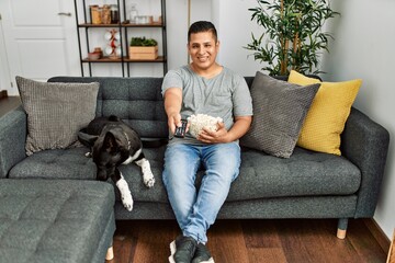 Young hispanic man watching movie sitting on the sofa with dog at home.