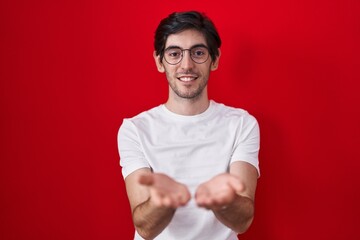 Naklejka premium Young hispanic man standing over red background smiling with hands palms together receiving or giving gesture. hold and protection