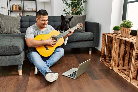 Young hispanic man having onlinr classical guitar lesson sitting on the floor with dog at home.