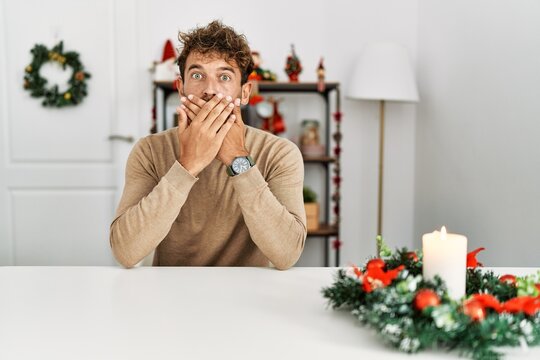 Young handsome man with beard sitting on the table by christmas decoration shocked covering mouth with hands for mistake. secret concept.