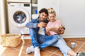 Young couple smiling happy reading book and using smartphone while doing laundry at home.