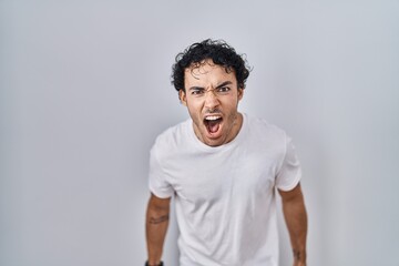 Hispanic man standing over isolated background angry and mad screaming frustrated and furious,...