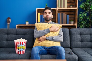 Hispanic man with beard eating popcorn watching a movie at home looking at the camera blowing a...