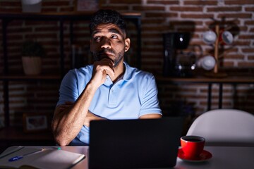 Fototapeta na wymiar Hispanic man with beard using laptop at night with hand on chin thinking about question, pensive expression. smiling with thoughtful face. doubt concept.
