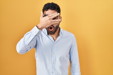 Fototapeta premium Hispanic man with beard standing over yellow background peeking in shock covering face and eyes with hand, looking through fingers with embarrassed expression.