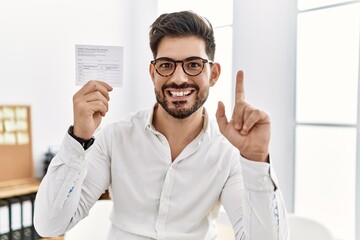 Young man with beard holding covid record card surprised with an idea or question pointing finger with happy face, number one