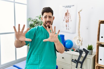 Young man with beard working at pain recovery clinic afraid and terrified with fear expression stop gesture with hands, shouting in shock. panic concept.