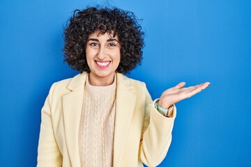 Young brunette woman with curly hair standing over blue background smiling cheerful presenting and pointing with palm of hand looking at the camera.