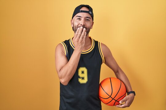 Middle age bald man holding basketball ball over yellow background laughing and embarrassed giggle covering mouth with hands, gossip and scandal concept