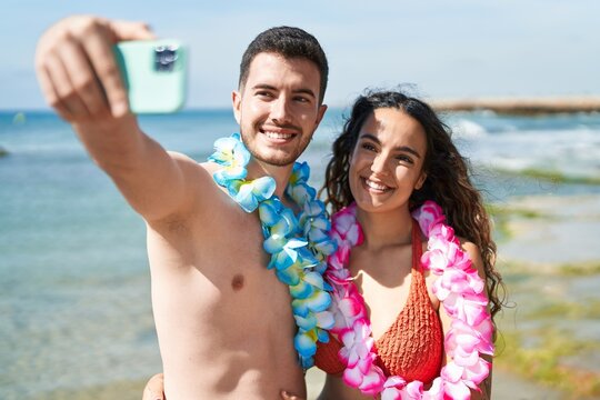 Young hispanic couple tourists wearing hawaiian lei make selfie by smartphone at seaside