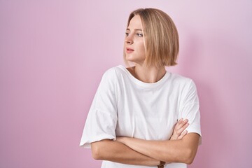 Young caucasian woman standing over pink background looking to the side with arms crossed convinced and confident