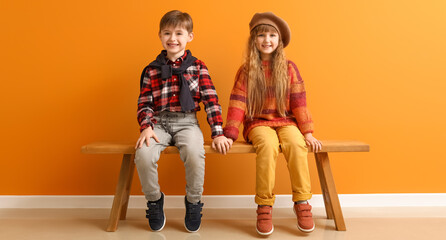 Cute little children in autumn clothes sitting on bench near orange wall
