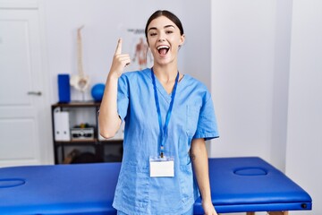 Young hispanic woman wearing physiotherapist uniform standing at clinic pointing finger up with...