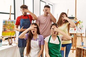 Group of five hispanic artists at art studio peeking in shock covering face and eyes with hand, looking through fingers with embarrassed expression.