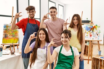 Group of five hispanic artists at art studio smiling and confident gesturing with hand doing small size sign with fingers looking and the camera. measure concept.