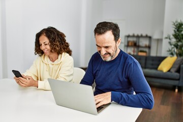 Middle age hispanic couple smiling happy using laptop and smartphone at home.