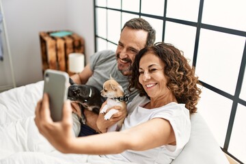 Middle age hispanic couple smiling happy making selfie by the smartphone. Lying on the bed with dogs at home.