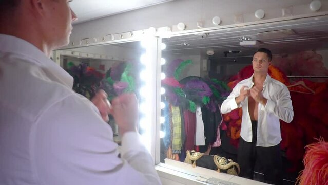 Handsome young man in dressing room gets dressed before performance,naked waist with tanned torso,pendant around his neck,puts on white shirt,looking at his mirror reflection.Stage costumes around.