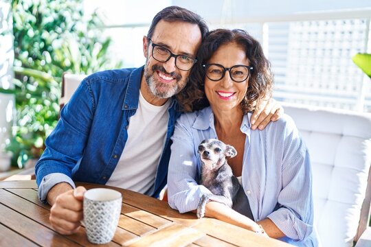 Middle age hispanic couple hugging each other sitting on table with dog at terrace
