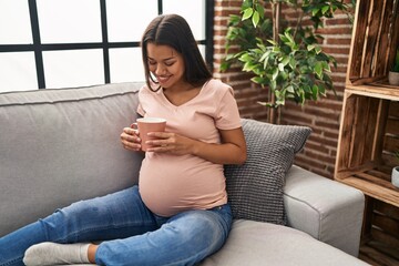 Young latin woman pregnant drinking coffee sitting on sofa at home