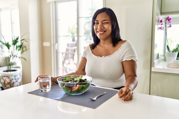 Young hispanic woman eating healthy salad at home winking looking at the camera with sexy...