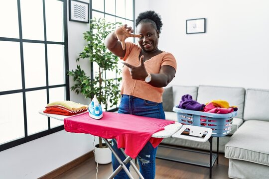 Young african woman ironing clothes at home smiling making frame with hands and fingers with happy face. creativity and photography concept.