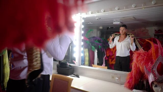 Pasadoble dancer in white shirt in dressing room in front of mirror looks at his reflection,slowly rotating throws bullfighter's vest over shoulders.Costumes,wigs and feathers,head mannequins around.