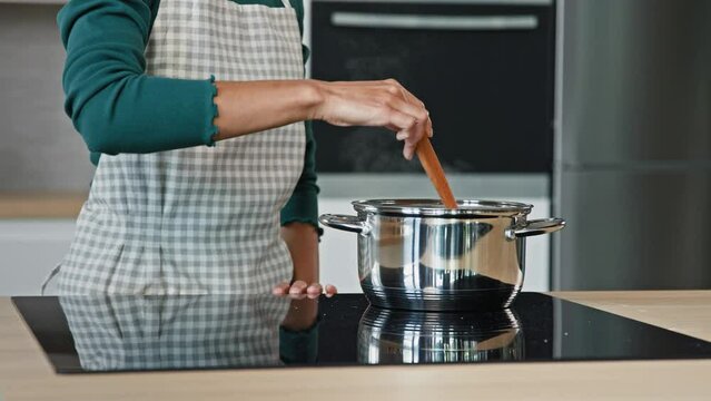Unknown housewife mistress maid wife mom wears apron prepare food on kitchen surface electric oven cooking aromatic dish boil off soup aromatic borsch by cookery recipe stir porridge with wooden spoon