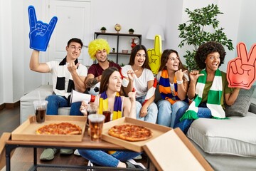 Group of young people wearing team scarf cheering football game pointing thumb up to the side smiling happy with open mouth