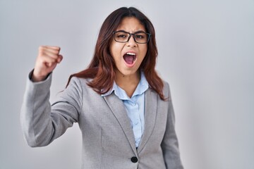 Hispanic young business woman wearing glasses angry and mad raising fist frustrated and furious...